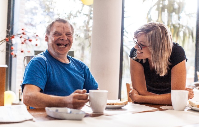 Een man een vrouw zitten lachend aan tafel. Beiden een beker en een bord met boterham voor zich. De man links lacht voluit richting camera en draagt een blauw t-shirt. De vrouw leunt met onderarmen op tafel, draagt een bril en zwart t-shirt en lacht richting de man. Op de achtergrond licht dat door grote ramen binnenvalt.