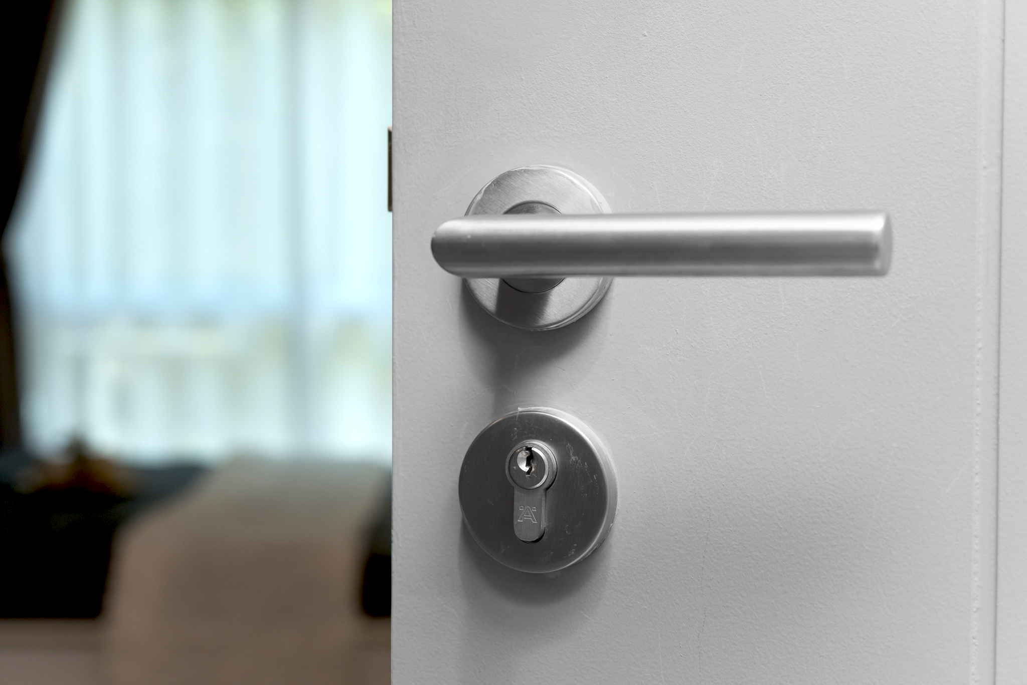 Witte deur met metalen deurklink en slot. De deur staat op een kier. Achter de kier is een raam met daglicht en een opgemaakt bed te zien.